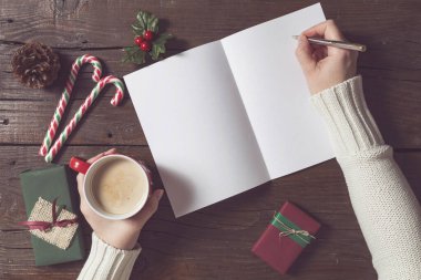Kadın ellerinin bir fincan kahve tutup Noel Baba 'ya mektup yazışının en üst görüntüsü. Noel hediyeleri ve süslemeler mektubun yanındaki ahşap masaya konmuş.