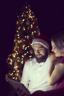 Güzel dekore edilmiş bir Noel ağacının yanında oturan aşık çift tablet bilgisayardaki bazı fotoğraflara bakıyor. Adama odaklan.