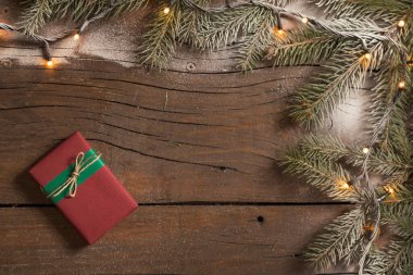 Karlı çam dallarından yapılmış bir çerçeve ve Noel ışıklarından yapılmış güzel paketlenmiş bir hediye kutusu ahşap masaya yerleştirilmiş.