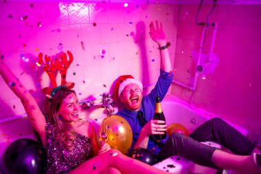 Yeni yıl partisinde eğlenen, şampanya içen, balon ve konfetiyle dolu küvette otururken şarkı söyleyen ve dans eden neşeli genç çiftin yüksek açılı görüntüsü.