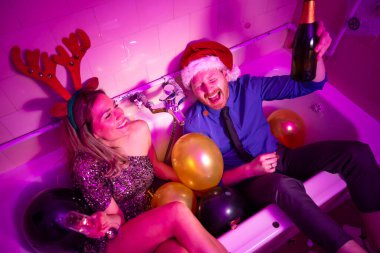 Yeni yıl partisinde eğlenen, şampanya içen, balonlarla dolu küvette otururken şarkı söyleyip dans eden neşeli genç çiftin yüksek açılı görüntüsü.