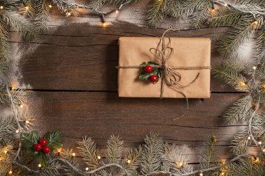 Karlı çam dallarından yapılmış bir çerçeve ve Noel ışıklarından yapılmış güzel paketlenmiş bir hediye kutusu ahşap masaya yerleştirilmiş.