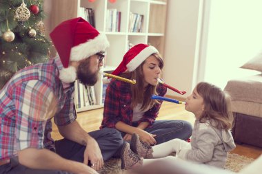 Güzel dekore edilmiş bir Noel ağacının yanında Noel Baba şapkası takan ve parti düdüğü çalan güzel bir aile. Annesine odaklan.