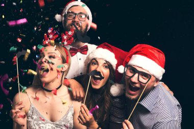 Noel Baba şapkası takan bir grup genç arkadaş yılbaşı partisinde eğleniyor, maskeli fotoğraflara poz veriyorlar.