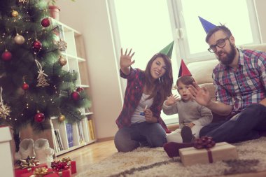 Güzel, genç bir aile Noel tatili için birlikte eğleniyor, oturma odasında yerde oturuyor, güzel dekore edilmiş bir Noel ağacının yanında, gülümsüyor ve el sallıyorlar. Bebeğe odaklan.