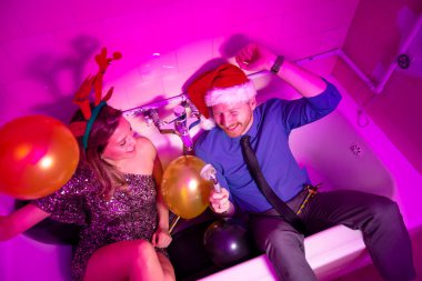 Yeni yıl partisinde, balonlarla dolu bir küvette oturmuş, mikrofon olarak karaoke yapan neşeli çiftin yüksek açılı görüntüsü. 