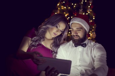 Güzel dekore edilmiş bir Noel ağacının yanında oturan aşık çift tablet bilgisayardaki bazı fotoğraflara bakıyor. Kıza odaklan.