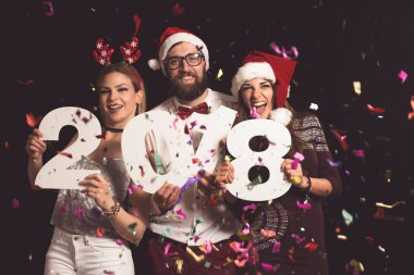 Üç genç arkadaş yeni yıl partisinde eğleniyor, ellerinde 2018 numaralı karton kutular var.