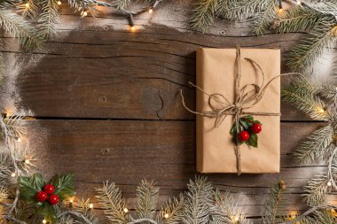 Karlı çam dallarından yapılmış bir çerçeve ve Noel ışıklarından yapılmış güzel paketlenmiş bir hediye kutusu ahşap masaya yerleştirilmiş.