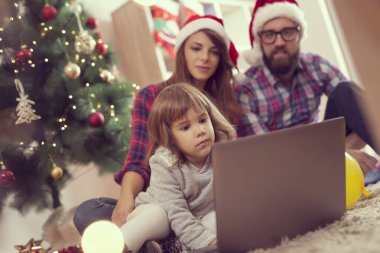 Genç ve güzel bir aile oturma odasının zemininde güzel dekore edilmiş bir Noel ağacının yanında oturmuş dizüstü bilgisayarında çizgi film seyrediyor. Bebeğe odaklan.