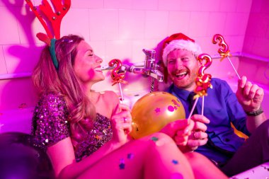 Birbirine aşık bir çift Noel Baba şapkası ve boynuzlarıyla balonlarla dolu bir küvette oturuyor ve yeni yıl partisinde eğleniyorlar, ellerinde 2023 numaralı balonlar var.