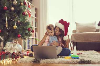 Anne ve kızı bir Noel ağacının yanında oturmuş dizüstü bilgisayarda çizgi film izliyor ve eğleniyorlar. Bebeğe odaklan.
