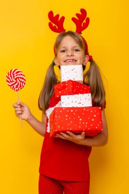 Güzel küçük kız elinde bir sürü Noel hediyesi ve büyük kırmızı beyaz lolipop ile sarı renkli arka planda izole edilmiş fotokopi alanı ile.