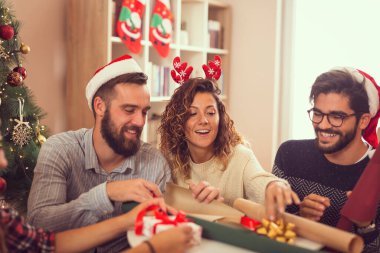 Bir grup genç arkadaş Noel ağacının yanında oturuyor, Noel hediyelerini paketliyor ve eğleniyorlar.