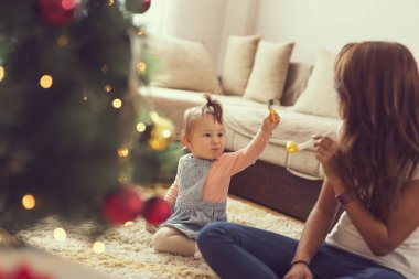 Güzel, genç bir anne ve kız Noel ağacının yanında oturmuş Noel sabahı parti ıslıkları çalıyorlar.