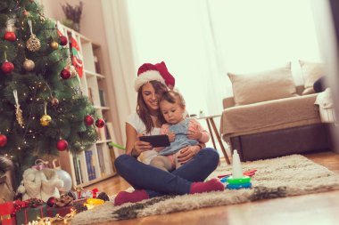 Anne ve kızı bir Noel ağacının yanında oturmuş akıllı bir telefonla çizgi film izliyorlar.