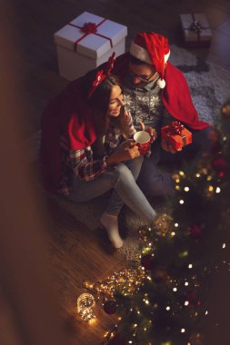 Güzel dekore edilmiş bir Noel ağacının yanında oturan, yumuşak kırmızı battaniyeyle örtülü, sıcak çay içen ve hediyelerini veren çiftin yüksek açılı görüntüsü.