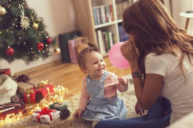 Güzel, genç bir anne ve kız Noel ağacının yanında oturup Noel sabahı balon şişirip oynuyorlar.