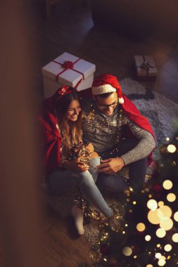 Güzel dekore edilmiş bir Noel ağacının yanında oturan, yumuşak kırmızı battaniyeyle kaplanmış, Noel arifesi sihriyle sarılıp eğlenen çiftin yüksek açılı görüntüsü. 