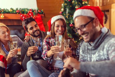 Bir grup arkadaş, güzel dekore edilmiş bir Noel ağacının yanında oturmuş bardaklara şampanya dolduruyorlar ve kadeh kaldırıyorlar.