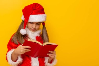 Noel Baba gibi giyinmiş çocuk kitap okuyor, sarı renkli arka planda kopya alanı ile izole edilmiş.