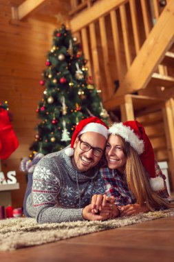 Güzel genç çift aşık güzel bir şömine ve A yanındaki yere yatarken Santa şapka giymiş Noel ağacı ve zevk Noel sabahı dekore edilmiştir