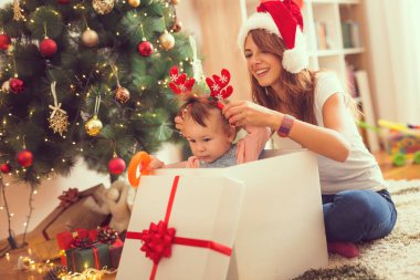 Genç anne bir Noel ağacının yanında yerde oturuyor ve hediye kutusundan atlayan küçük kızıyla oynuyor. Bebeğe odaklan.