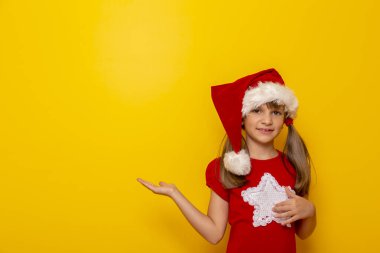 Noel Baba şapkası takan çocuk yanındaki boşluğu işaret ediyor ve sarı renkli arka planda izole bir şekilde gülümsüyor.