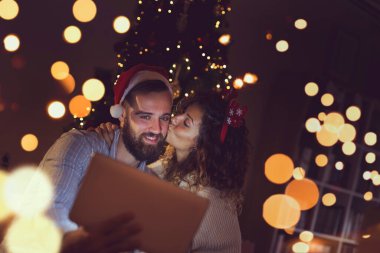 Birbirine aşık bir çift Noel ağacının yanında oturuyor, Noel Baba şapkası takıyor ve tablet bilgisayardaki eski fotoğraflara bakarak eğleniyor..