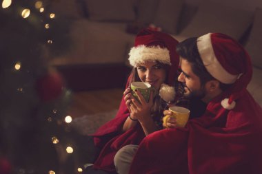 Birbirine aşık bir çift Noel ağacının yanında oturuyor, Noel Baba 'nın şapkalarını takıyor, sıcak çay içiyor ve birlikte romantik anların tadını çıkarıyor.