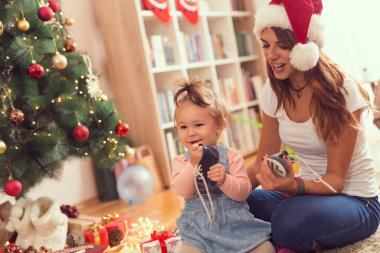 Güzel, genç bir anne ve kız güzel dekore edilmiş bir Noel ağacının yanında oturup Noel sabahı eğleniyorlar.