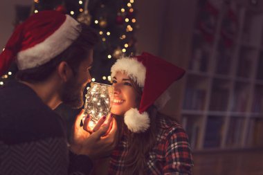 Güzel bir çift Noel ağacının yanında, oturma odasında oturmuş Noel ışıklarıyla dolu bir kavanozu tutuyorlar.