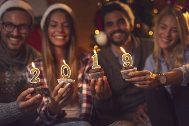 Bir grup genç arkadaş Noel ağacının yanında Noel Baba 'nın şapkalarını takıp üzerinde 2019 mum olan kekleri tutuyorlar. Mumlara odaklan.