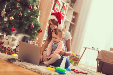 Anne ve kızı bir Noel ağacının yanında oturmuş dizüstü bilgisayarda çizgi film izliyor ve eğleniyorlar. Bebeğe odaklan.