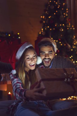 Aşık genç bir çift şöminenin yanında oturuyor ve güzel dekore edilmiş bir Noel ağacı, hediyeleri açıyor ve Noel arifesinde eğleniyorlar. Kıza odaklan.