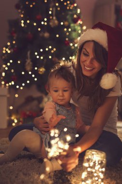 Güzel, genç bir anne ve kız Noel ağacının yanında oturmuş maytap ve yılbaşı ışıklarıyla oynuyorlar. Bebeğe odaklan.