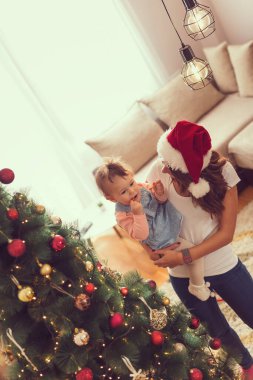 Güzel, genç bir anne ve kız Noel 'de evde birlikte vakit geçiriyorlar, anne Noel Baba şapkası takıyor ve bebeği Noel ağacının yanında tutuyor.