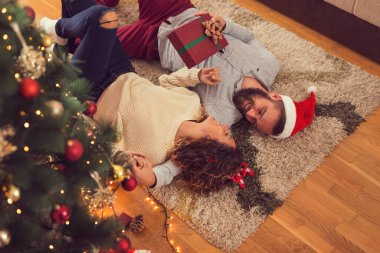 Güzel, genç bir çiftin, güzel dekore edilmiş bir Noel ağacının yanında, oturma odasının zeminine uzanmış, birbirlerine Noel hediyelerini verirken yüksek açılı görüntüsü.