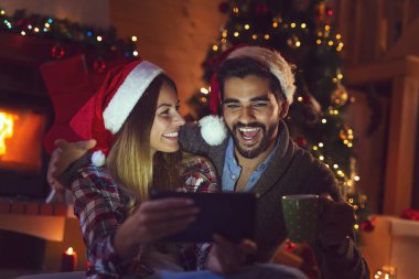Aşık bir çift Noel ağacı ve şöminenin yanında oturuyor, Noel Baba şapkaları takıyor ve tablet bilgisayardaki eski fotoğraflara bakarak eğleniyor.