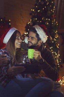 Aşık bir çift Noel ağacı ve şöminenin yanında oturuyor, Noel Baba şapkaları takıyor ve tablet bilgisayardaki eski fotoğraflara bakarak eğleniyor. Adama odaklan.