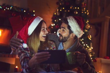 Aşık bir çift Noel ağacı ve şöminenin yanında oturuyor, Noel Baba şapkaları takıyor ve tablet bilgisayardaki eski fotoğraflara bakarak eğleniyor.