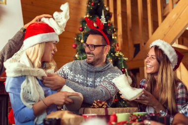 Güzel dekore edilmiş bir Noel ağacının yanında oturan bir grup genç arkadaş, Noel hediyelerini değiş tokuş edip eğleniyorlar. Ortadaki adama odaklan.