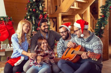 Bir grup arkadaş güzel dekore edilmiş bir Noel ağacının yanında oturmuş gitar çalıyor, Noel şarkıları söylüyor ve şarap içiyor.