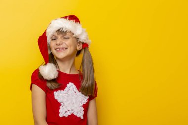 Noel Baba şapkası takan güzel bir kızın portresi gülümseyen ve göz kırpan, sarı renkli arka planda izole edilmiş, fotokopi alanı olan