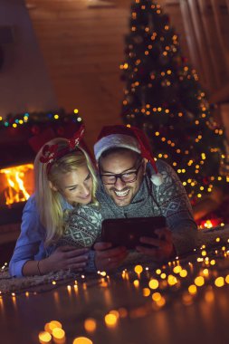 Birbirine aşık bir çift Noel ağacının yanında oturuyor, Noel Baba şapkası takıyor ve tablet bilgisayardaki eski fotoğraflara bakarak eğleniyor.