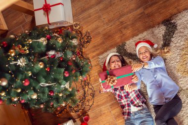 İki güzel kadının oturma odasının zemininde uzanmış Noel ağacının yanında güzel paketlenmiş Noel hediyelerini tutarken görüntüsü.