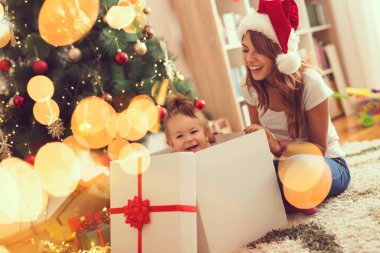 Genç anne bir Noel ağacının yanında oturmuş hediye kutusundan atlayan küçük kızıyla oynuyor.