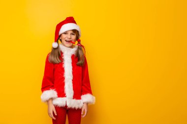 Noel Baba kostümü giymiş güzel bir kızın portresi, parti düdüğü çalarken eğleniyor, sarı renkli arka planda izole edilmiş, fotokopi alanı olan.