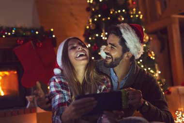 Aşık bir çift Noel ağacı ve şöminenin yanında oturuyor, Noel Baba şapkaları takıyor ve tablet bilgisayardaki eski fotoğraflara bakarak eğleniyor.