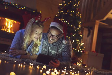 Birbirine aşık bir çift Noel ağacının yanında oturuyor, Noel Baba 'nın şapkalarını takıyor ve tablet bilgisayardaki eski fotoğraflara bakarak eğleniyor. Adama odaklan.
